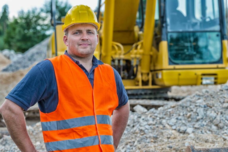 Een bouwvakker met een oranje veiligheidsvest en gele helm staat voor een gele graafmachine op een rotsachtige bouwplaats.