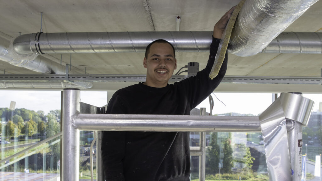 A man on a scaffold looking at the camera. His left hand is resting on a pipe covered with insulation.
